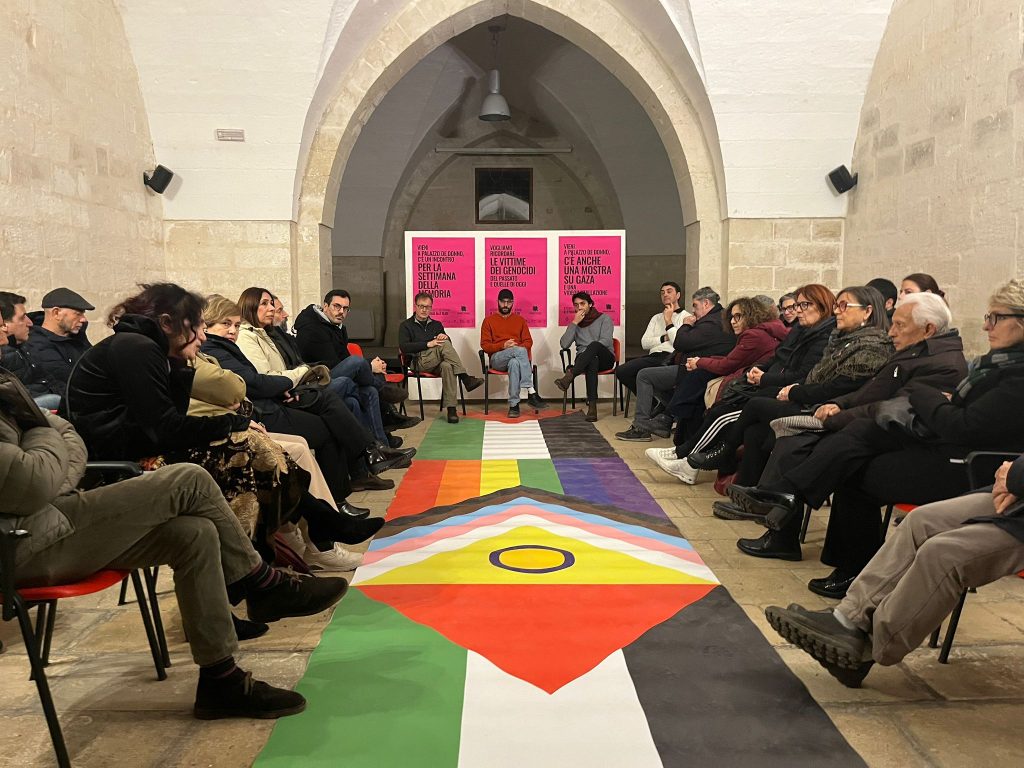 platea di persone discute sul tema della guerra in Palestina, sala allestita con una bandiera palestinese e della comunità lgbtiqa+