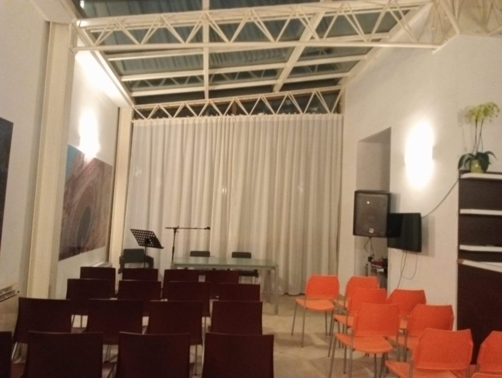 Immagine della sala polifunzionale dell'Ostello con una grande vetrata coperta da una tenda decorativa. In primo piano, un banco per i relatori e diverse sedute disposte frontalmente per gli interlocutori. L’ambiente è luminoso, ordinato e predisposto per eventi pubblici o incontri formativi.