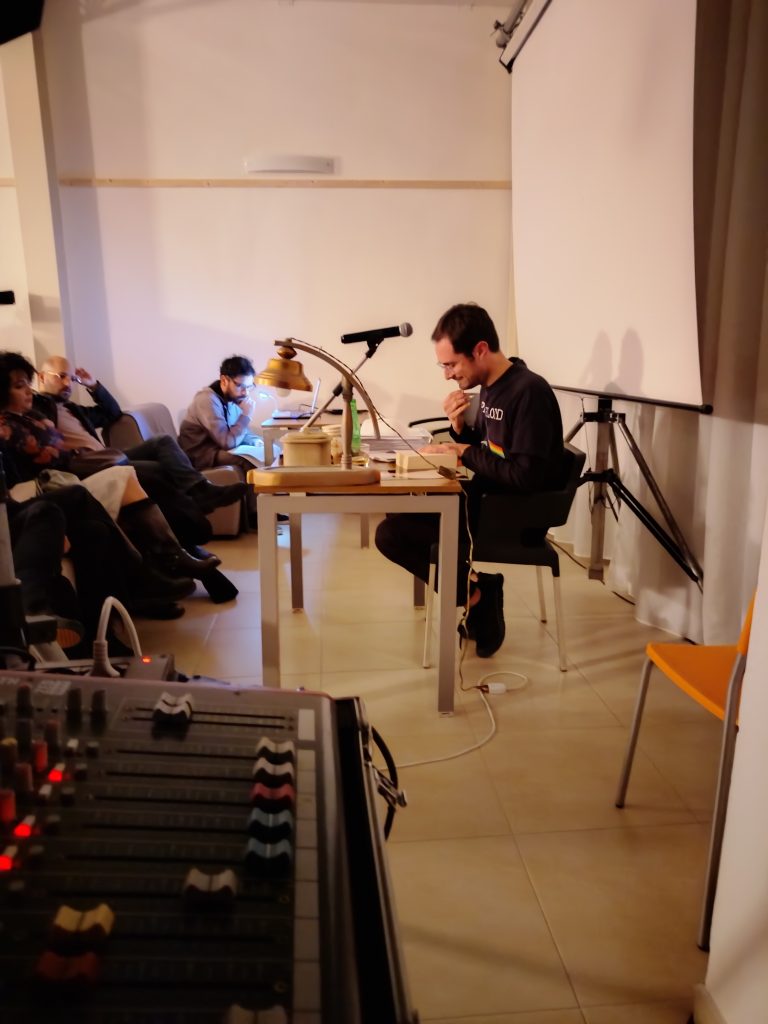 Lorenzo Di Lauro seduto al tavolino durante lo spettacolo “I Fantasmi di Floyd” all’Ostello Novello, intento a raccontare la storia dei Pink Floyd.