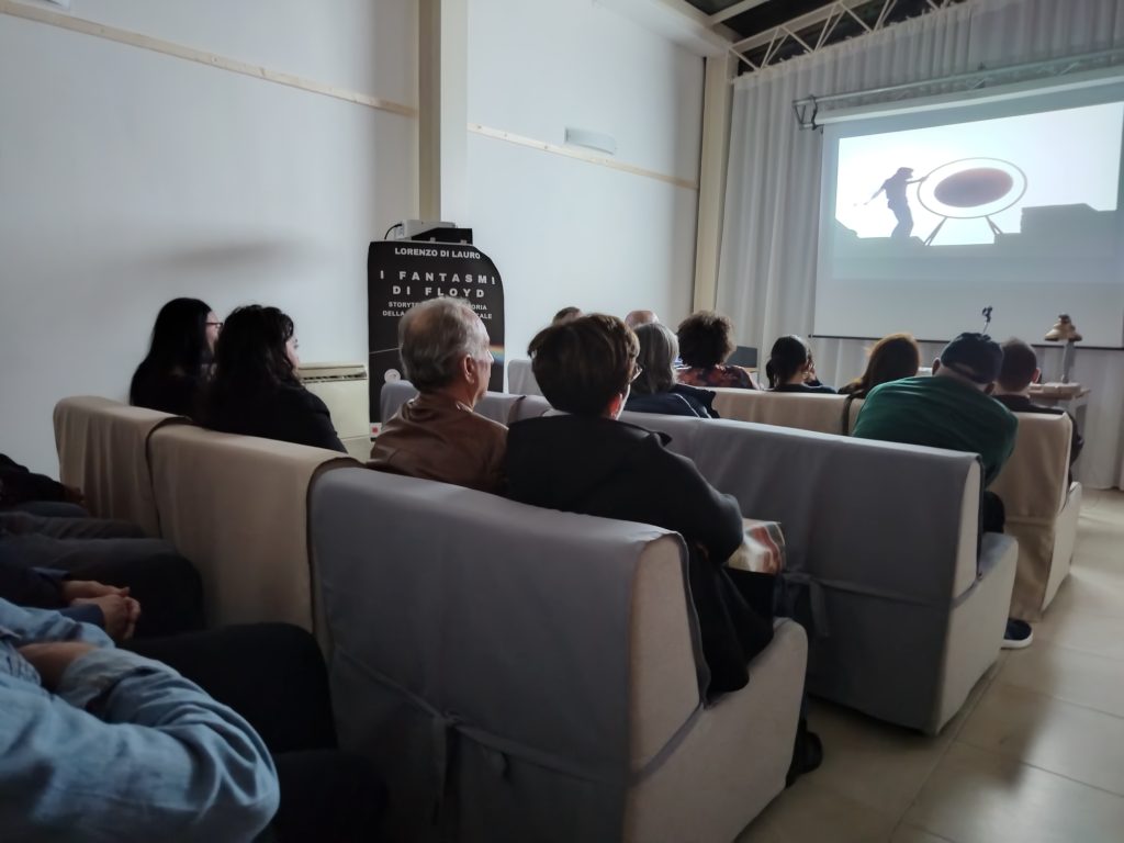 Pubblico seduto all’Ostello Novello, concentrato sulla visione di contenuti multimediali sui Pink Floyd dopo lo spettacolo “I Fantasmi di Floyd”.