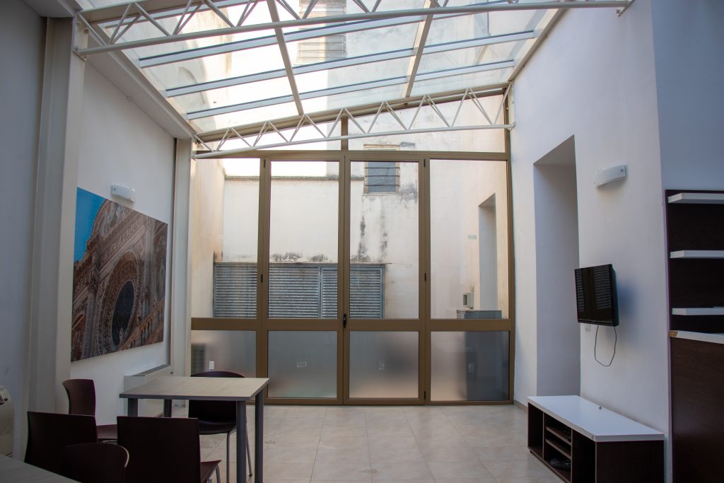 Sala vuota e minimalista con pavimento chiaro e pareti neutre; sullo sfondo, una porta vetrata lascia intravedere la luce naturale proveniente dall’esterno.