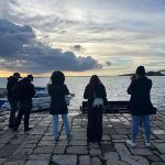 Sei persone, studenti di un corso di fotografia, sono rivolte verso un lago o il mare con cielo nuvoloso al tramonto, scattando foto. Sono su un molo pavimentato in pietra.