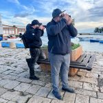 Due uomini, uno in primo piano con un berretto e l'altro leggermente dietro, stanno scattando foto su un molo pavimentato in pietra. Entrambi tengono una fotocamera (o un telefono) davanti al viso. Lo sfondo mostra un porto con piccole barche ormeggiate, una panchina di legno e un cielo parzialmente nuvoloso sopra l'acqua.