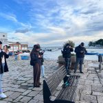 Quattro persone, due donne e due uomini, si trovano su una banchina lastricata, tutti intenti a fotografare o usare i loro dispositivi con vista su un piccolo porto con barche ormeggiate e un cielo parzialmente nuvoloso all'imbrunire.