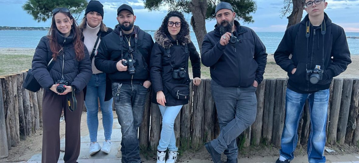 Gruppo di sei studenti di fotografia in piedi all'aperto, in posa per una foto di gruppo davanti a una staccionata di legno su una spiaggia. Hanno tutti indosso giacche scure e tengono fotocamere digitali reflex al collo o tra le mani. Il paesaggio retrostante include alberi e il mare in una giornata nuvolosa.