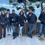 Gruppo di sei studenti di fotografia in piedi all'aperto, in posa per una foto di gruppo davanti a una staccionata di legno su una spiaggia. Hanno tutti indosso giacche scure e tengono fotocamere digitali reflex al collo o tra le mani. Il paesaggio retrostante include alberi e il mare in una giornata nuvolosa.