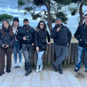 Gruppo di sei studenti di fotografia in piedi all'aperto, in posa per una foto di gruppo davanti a una staccionata di legno su una spiaggia. Hanno tutti indosso giacche scure e tengono fotocamere digitali reflex al collo o tra le mani. Il paesaggio retrostante include alberi e il mare in una giornata nuvolosa.