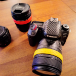 Una fotocamera reflex digitale Canon EOS 77D con un obiettivo 18-135mm, affiancata da altri due obiettivi chiusi con tappi, posati su un tavolo di legno