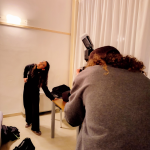 Uno studente, in primo piano e di spalle, indossa un maglione grigio e sta fotografando l'altro studente, vestito di nero, che posa sorridendo con il busto inclinato all'indietro. La scena è illuminata da un flash sulla fotocamera e mostra sullo sfondo tende bianche e un tavolo di lavoro.