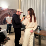 Un uomo con un cappellino e un cappuccio nero parla con una donna che indossa un maglione e pantaloni chiari, tenendo in mano un obiettivo fotografico, in un ambiente interno con tende bianche e altre persone sedute.