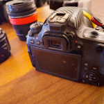 Una fotocamera reflex digitale Canon EOS 77D con un obiettivo 18-135mm, affiancata da altri due obiettivi chiusi con tappi, posati su un tavolo di legno