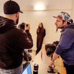 Due fotografi in primo piano, di cui uno con cappellino, sono impegnati in un'esercitazione pratica. Uno sta inquadrando un soggetto femminile posizionato contro una parete bianca. L'altro fotografo (con cappello multicolore) sta parlando o dando indicazioni, appoggiato a un tavolo di legno su cui sono presenti attrezzature fotografiche, uno smartphone e un computer portatile acceso in primo piano.