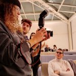 Primo piano su un gruppo di studenti di fotografia in una lezione pratica. Una persona con capelli ricci e un berretto tiene una fotocamera reflex con un flash montato sulla slitta e la guarda mentre riceve istruzioni dall'istruttore Mattia accanto a lei. Sullo sfondo, in primo piano sulla destra, una donna in maglione a collo alto è seduta e osserva la scena. Altri partecipanti si intravedono in fondo alla stanza.