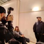 Tre persone in un ambiente interno minimalista. Una persona, ripresa di spalle in primo piano, tiene una fotocamera con flash rivolta verso due soggetti. Un soggetto è seduto e guarda verso l'obiettivo, l'altro è in piedi a destra e guarda in avanti. Scatto durante un corso di fotografia.