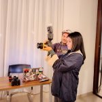 Due persone, un uomo (l'istruttore) e una donna (la studentessa), si trovano in un interno luminoso. L'istruttore, con un berretto, sta mostrando alla studentessa come tenere e usare una fotocamera reflex con un flash esterno montato sopra, durante una lezione pratica di fotografia. Sul tavolo di legno accanto a loro ci sono un computer portatile con molti adesivi, un'altra fotocamera e un telecomando.