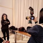 Una studentessa in primo piano, vista di spalle, sta scattando una foto usando una fotocamera reflex con un flash esterno montato. Il suo soggetto è un'altra studentessa, con i capelli ricci e gli occhiali, che posa con le braccia incrociate sullo sfondo, di fronte a una tenda bianca. Un momento pratico di ritratto durante un corso di fotografia.