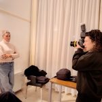 Donna con capelli ricci scuri che fotografa con una reflex e flash, mentre un'altra donna bionda con maglione a collo alto e jeans è in piedi sullo sfondo con le braccia conserte.