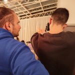 Due uomini durante il corso di fotografia. L'uomo in felpa blu (Mattia) ha i capelli raccolti in uno chignon e una barba, ed è ritratto di profilo mentre osserva l'uomo accanto a lui, visto di schiena, che tiene una fotocamera e sembra stia scattando una foto o esercitandosi. Si intravede un soffitto con travature e una tenda chiara sullo sfondo.