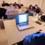 Istantanea di una lezione frontale in aula. Un gruppo di studenti è seduto su un divano dietro un tavolo di legno chiaro. La maggior parte degli studenti guarda un computer portatile aperto in primo piano sul tavolo, che mostra delle slide.