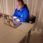 Ritratto di Mattia, l'istruttore del corso di fotografia, seduto a un tavolo di legno chiaro. Indossa una felpa blu acceso con cappuccio e sta lavorando concentrato al suo computer portatile, il cui coperchio è decorato con numerosi adesivi. Dietro di lui, una grande tenda bianca funge da sfondo neutro.