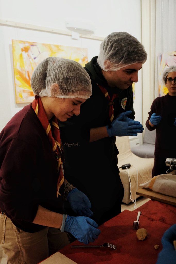 Due persone, dotate di dispositivi igienici, lavorano su un tavolo preparando materiali durante un laboratorio.